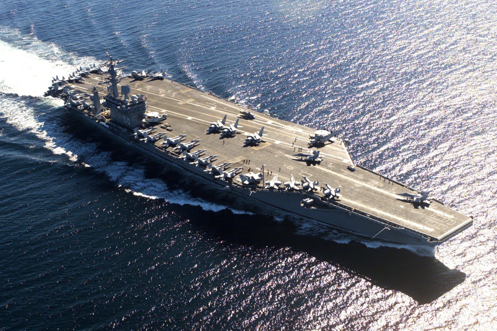 A US Navy aircraft carrier at sea seen from above, its flight deck lined with fighter jets