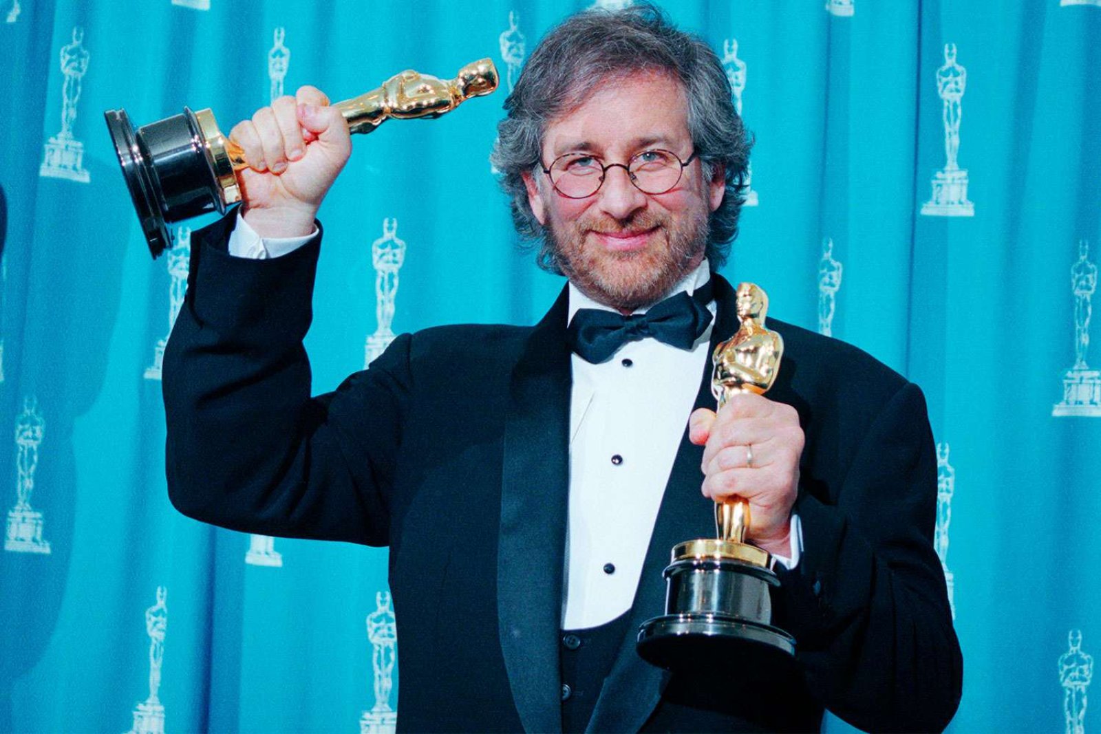 Steven Spielberg in a tuxedo and round glasses, smiling and holding up two Oscar statuettes