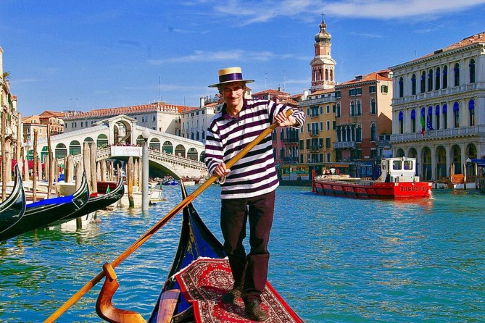 A gondolier in a straw hat and striped shirt rowing a gondola on the Grand Canal, with the Rialto Bridge behind him